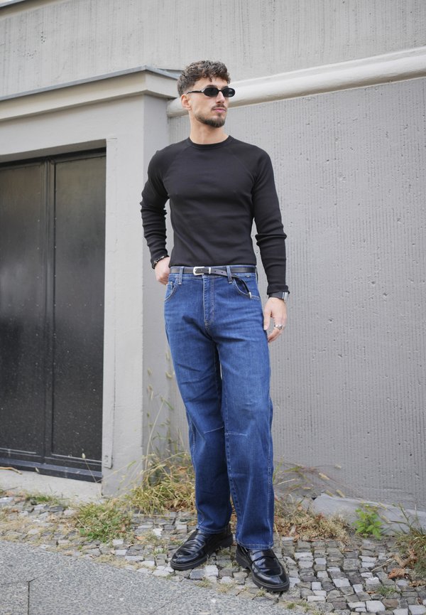 Hombre con camisa negra de manga larga, vaqueros azules, zapatos negros, gafas de sol, de pie en una acera de adoquines cerca de una pared gris de un edificio.