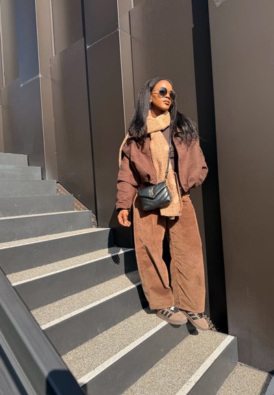 Mujer con conjunto de pana marrón, bufanda beige con estampado, gafas de sol y bolso negro cruzado, parada en unas escaleras al aire libre bajo la luz del sol.