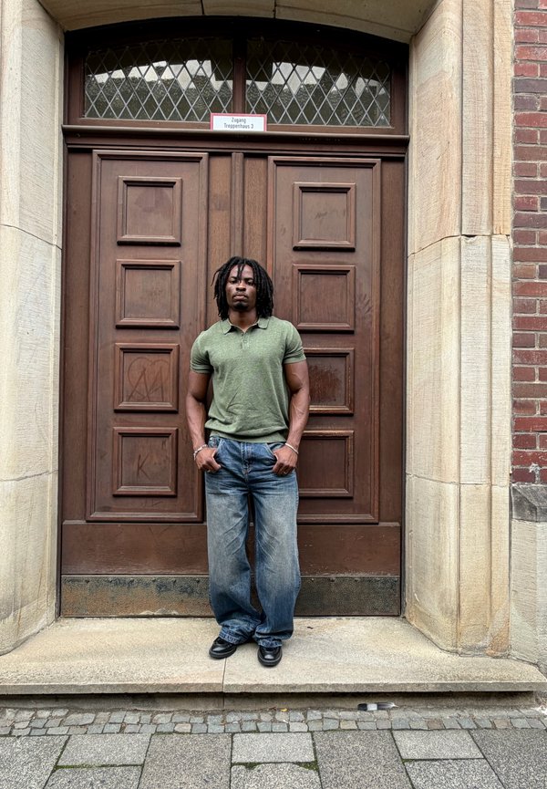 Homme avec des dreadlocks portant un polo vert et un jean bleu se tient devant de grandes portes doubles en bois avec un encadrement en pierre et en brique.