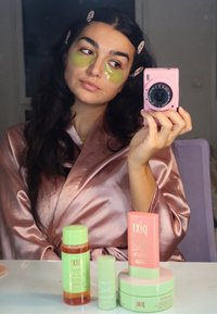 Ein rosa Satinbademantel umhüllt eine Person mit grünen Augenschutzmasken. Eine hellrosa Kamera spiegelt sich in der Hand wider. Hautpflegeprodukte in Grün und Pink sind auf einem Tisch angeordnet.