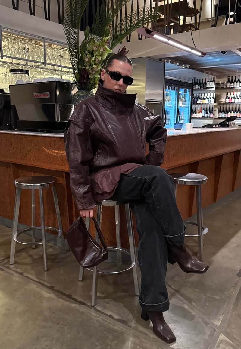 Giacca in pelle marrone con colletto alto, pantaloni in denim nero, borsa marrone e stivali con tacco, seduta a un bancone con bicchieri sullo sfondo.