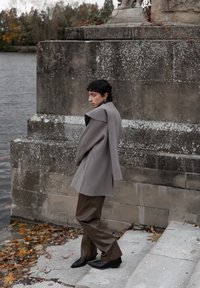 Person iført en grå frakke og brune bukser, stående på sten trapper ved siden af en stor, vejrslidt stenskulptur nær en vandkrop.