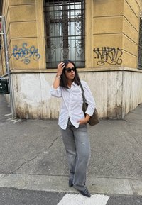 Jonge vrouw met zwarte zonnebril, witte shirt, grijze jeans en zwarte laarzen staat bij een gebouw met graffiti, terwijl ze een bruine tas vasthoudt.
