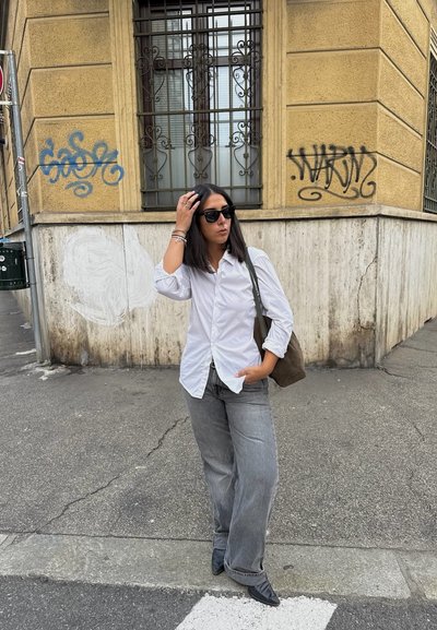 Joven mujer con gafas de sol negras, camiseta blanca, jeans grises y botas negras se encuentra junto a la esquina de un edificio marcado con graffiti, sosteniendo una bolsa marrón.