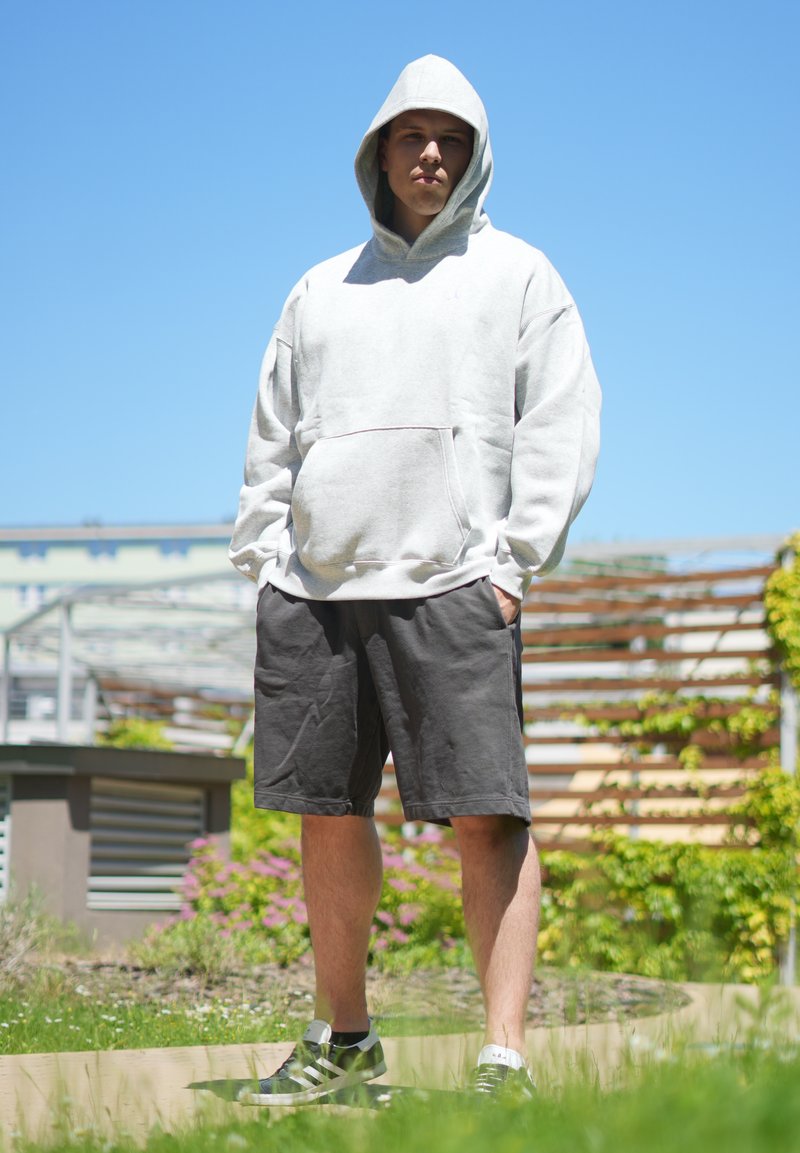 Junge Person trägt einen weissen Hoodie mit aufgesetzter Kapuze, schwarze Shorts und Sneakers, steht draussen mit den Händen in den Taschen unter klarem blauen Himmel.