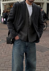 Blazer preto sobre um colete cinza de lã, camisa branca por baixo, combinado com jeans azuis largos. Pequena bolsa preta transversal, pavimento de tijolos visível.