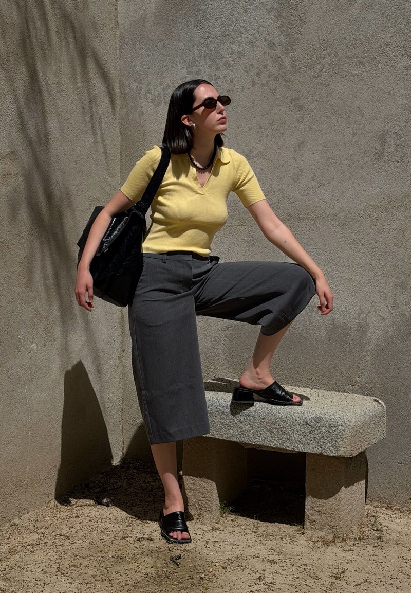 Femme portant un haut jaune et un pantalon gris court pose avec un pied sur un banc en pierre devant un mur texturé, portant un sac noir.
