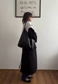 Femme portant un manteau noir et des lunettes de soleil, debout de profil avec un grand sac noir, sous un texte encadré indiquant « Hello MTV welcome to my crib »