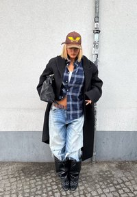 Personne portant une casquette brune, un manteau noir, une chemise à carreaux bleue, un jean large usé rentré dans des bottes noires, debout à côté d'un poteau couvert d'autocollants.