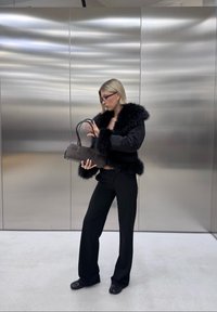 Sort jakke med faux pelskrave, sorte tætsiddende bukser og en mørk lædertaske. Baggrunden har glatte metalpaneler.