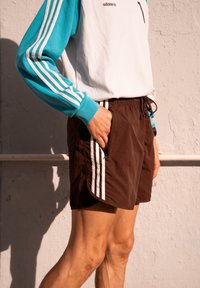 Shorts de sport marron avec des bandes blanches sur les côtés, dotés d'une taille à cordon de serrage et de poches. Associés à un haut à manches longues blanc et turquoise.