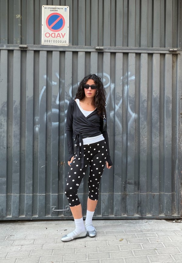 Mujer con gafas de sol, top negro cruzado, leggings de lunares, calcetines blancos y zapatos plateados, de pie frente a una puerta metálica gris con un cartel de prohibido aparcar.