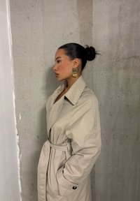 Trench coat bege com cintura cintuada, grandes lapelas e bolsos laterais. Brincos geométricos dourados complementam o conjunto contra um fundo cinza.