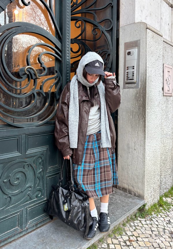 Femme portant une veste en cuir noire, une écharpe grise et une jupe à carreaux, debout près d'une porte en métal ornée à côté d'un panneau d'interphone.