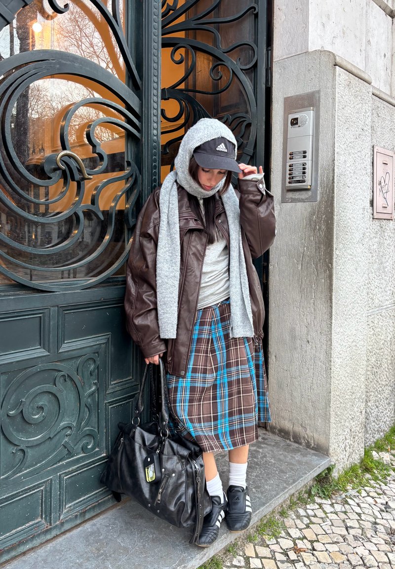 Kvinde iført mørk læderjakke, gråt tørklæde og ternet nederdel, stående ved en smukt udsmykket metal dør ved siden af en dørtelefon.