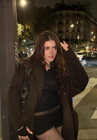 Mulher jovem com cabelo castanho longo, vestindo um fato preto e um casaco castanho oversized, parada num canto de rua da cidade à noite.