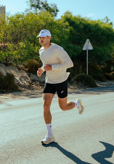 Camisa blanca de manga larga, pantalones cortos negros y zapatillas de correr con acentos amarillos. El corredor, con una gorra, trota por un camino iluminado por el sol. Vegetación verde cercana.