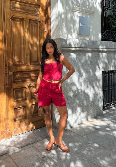 Top corto de lino rojo con pantalones cortos de tiro alto a juego, combinado con sandalias de punta abierta. Puerta de madera y pared de piedra en el fondo.