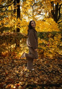 Joven sonriendo, posando con una pierna levantada en un camino cubierto de hojas, rodeado de árboles y arbustos amarillos de otoño.