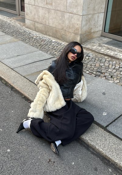 Chaqueta corta de cuero negro, abrigo de piel sintética crema, pantalones anchos negros, tacones puntiagudos negros y calcetines blancos, sentado en la acera.