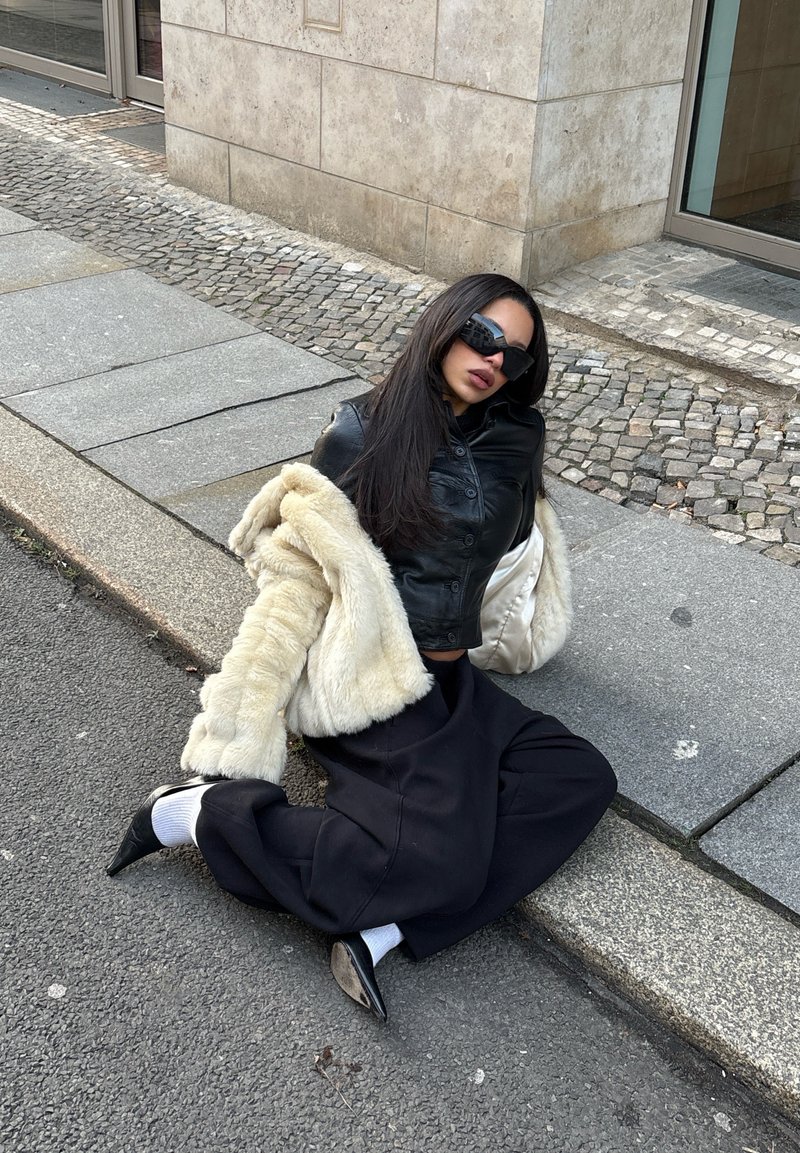 Veste courte en cuir noir, manteau en fausse fourrure crème, pantalon large noir, escarpins noirs pointus et chaussettes blanches, assis sur le trottoir.