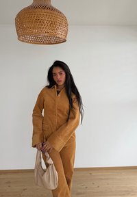 Femme debout à l'intérieur portant un tailleur en velours côtelé marron, tenant un sac à main beige, avec un grand abat-jour tissé suspendu au-dessus, devant un mur blanc.