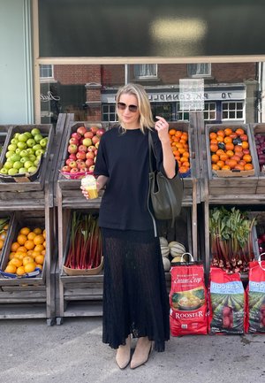 Camiseta negra oversized, falda larga de encaje negra, botas de tobillo beige. Sosteniendo un vaso transparente con bebida naranja. El fondo presenta puestos de frutas de madera.