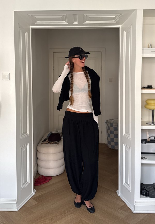Mujer con dos largas trenzas, llevando gorra negra, gafas de sol, top blanco transparente, pantalones negros y zapatos negros, de pie en un marco de puerta blanco.