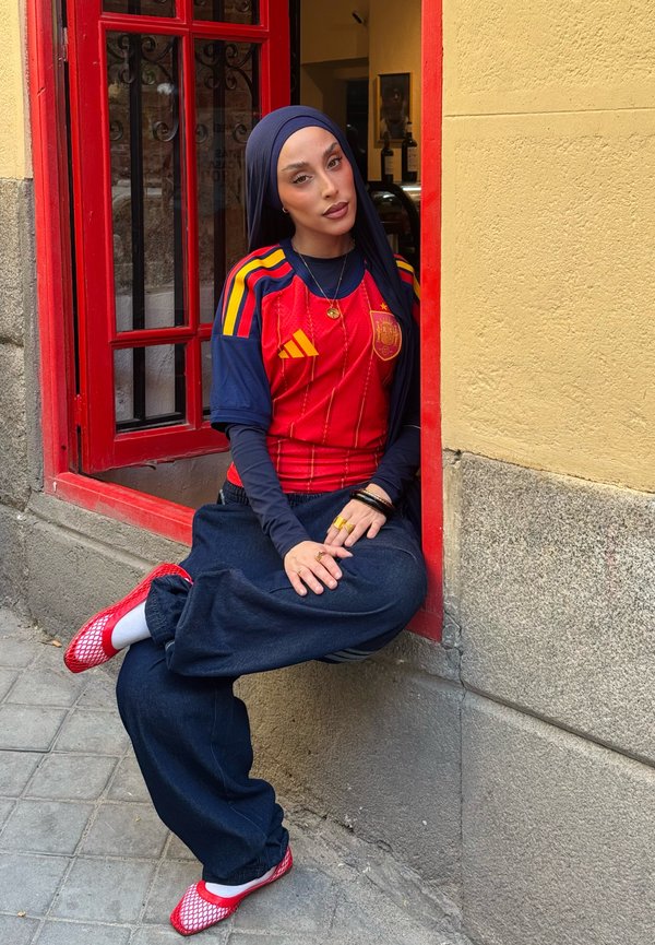 Jonge vrouw die een hijab en een voetbalshirt van Spanje draagt, zit ontspannen in een raam met een rode omlijsting op een hoek van een stadsstraat.