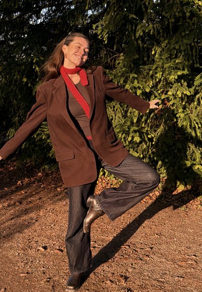 Mujer con blazer marrón, pantalones oscuros, bufanda roja y botas, de pie sobre una pierna con los brazos extendidos, sonriendo, frente a un follaje verde.