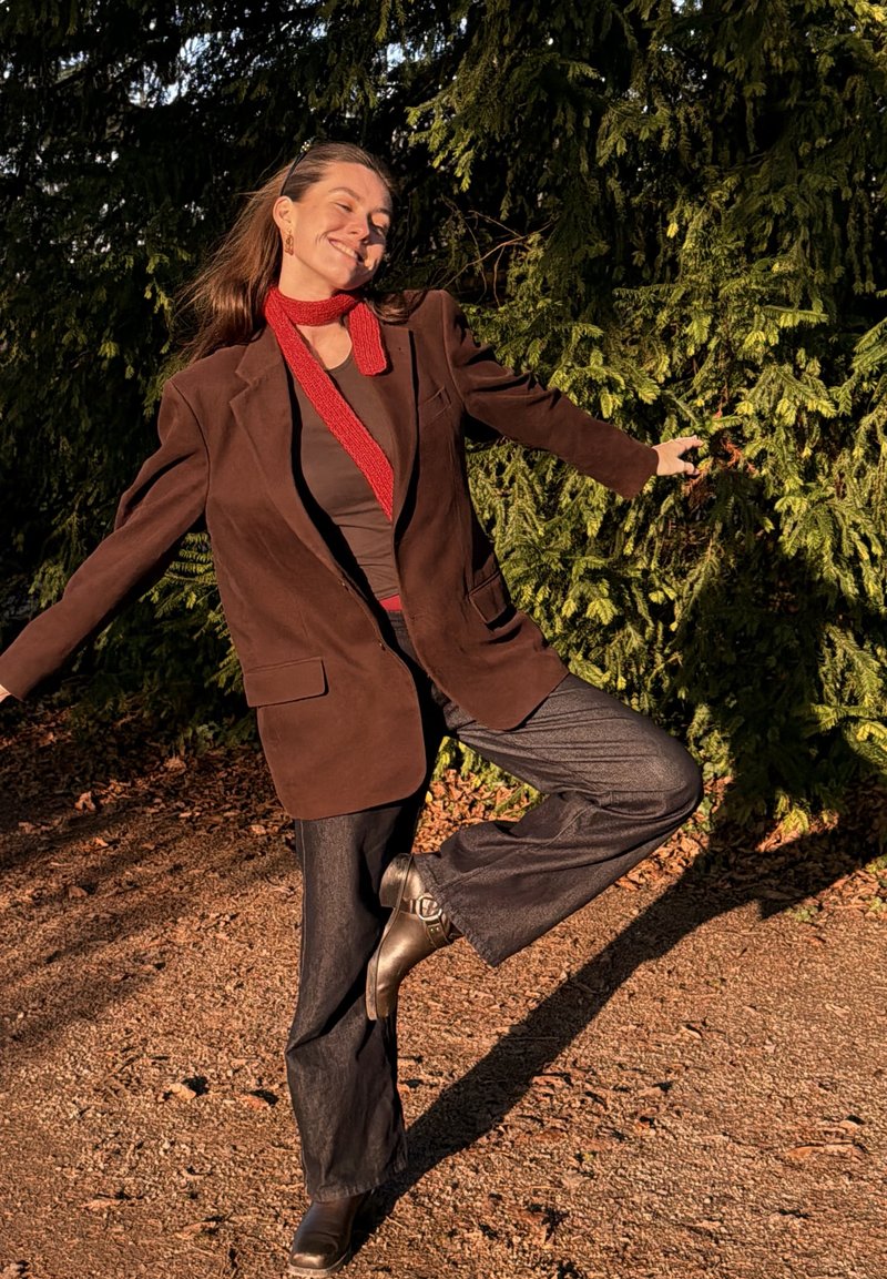 Femme portant un blazer marron, un pantalon foncé, une écharpe rouge et des bottes, debout sur une jambe avec les bras étendus, souriante, devant un feuillage vert.