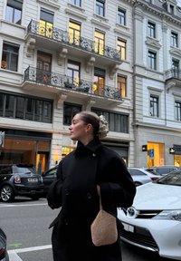 Manteau noir croisé en tissu texturé. Tient un petit sac à main clair orné de perles. Scène de rue avec des bâtiments et des voitures garées en arrière-plan.