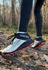 Scarpe da trail running GORE-TEX con tomaia prevalentemente bianca, dettagli neri e suola rosa. Presentano superficie testurizzata e lacci.