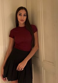 Μπορντώ εφαρμοστό μπλουζάκι συνδυασμένο με μια μαύρη πλισέ φούστα. Το ύφασμα είναι λείο με ορατές ραφές. Το σύνολο είναι μπροστά σε ένα ανοιχτόχρωμο τοίχο.