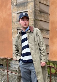 Light olive trench coat with a brown collar over a navy and white striped jumper. Grey trousers. Black baseball cap with a logo.