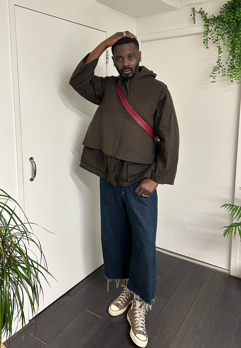Homme portant une veste marron superposée, un jean large bleu, des baskets hautes beige et un sac bandoulière rouge, debout à l'intérieur près de plantes.