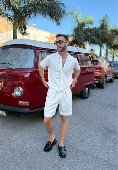 Camisa de punto blanca con cuello y botones, combinada con pantalones cortos blancos. Mocasines negros. Furgoneta roja vintage de fondo, con palmeras cerca.