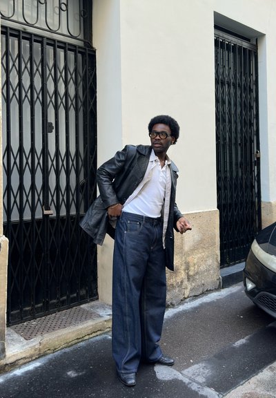 Hombre con chaqueta de cuero negra, camisa blanca y vaqueros anchos, de pie en la calle cerca de unas puertas metálicas negras y un coche estacionado.