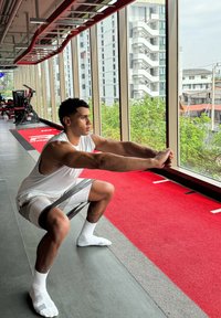 Athlète effectuant un squat dans une salle de sport, portant un débardeur blanc et un short gris. Il utilise une bande de résistance et se tient sur un sol gris.