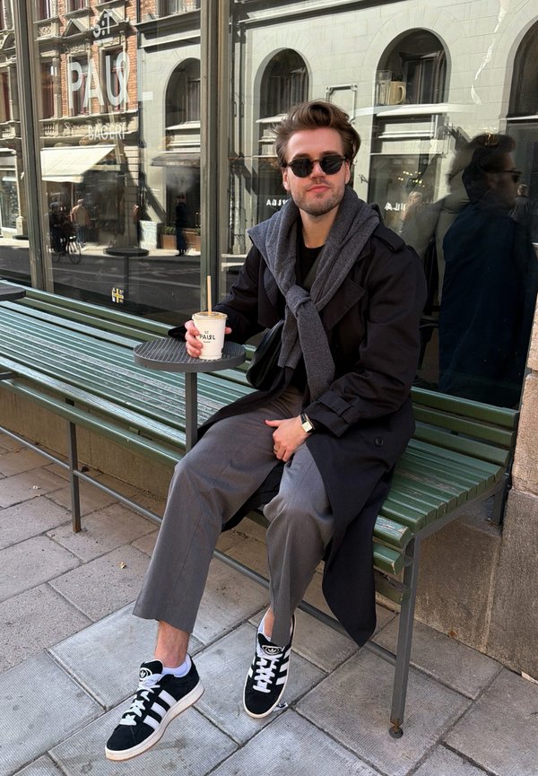 Uomo con occhiali da sole, trench coat e sneakers seduto su una panchina verde fuori da una panetteria, con in mano un bicchiere di caffè da asporto con cannuccia.