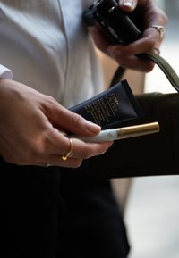 Estée Lauder Double Wear fondas, tamsiai mėlynas, su auksinėmis raidėmis, laikomas kartu su sidabriniu tušo vamzdeliu su auksiniu dangteliu, prieš tamsią rankinę.