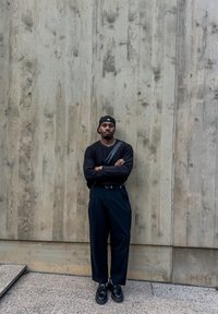 Maglione nero a maglia, pantaloni neri a vita alta, scarpe nere. In piedi contro un muro di legno texturizzato, braccia incrociate, indossa un cappellino.