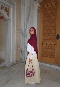 Maroon hijab, biały top z długim rękawem, beżowa spódnica. Nosi torbę w szachownicę i ma srebrne akcenty. Marmurowe tło i misternie rzeźbione drzwi.