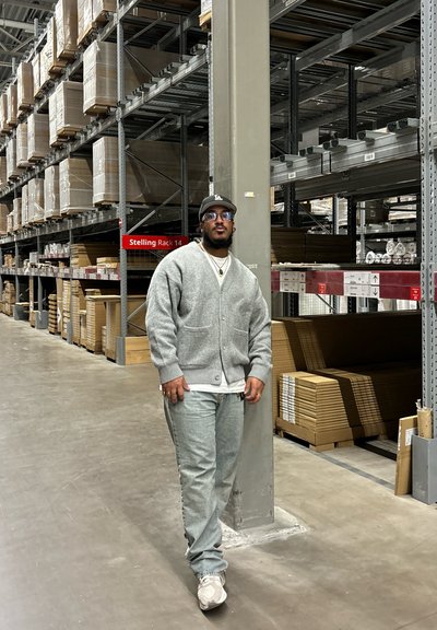 Cardigan gris con botones, camisa blanca y jeans de mezclilla azul claro. Fondo de almacén con estanterías apiladas con cajas y materiales.