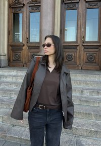 Καφέ δερμάτινο μπουφάν με φαρδιά μανίκια, τοποθετημένο πάνω από ένα σκούρο καφέ τοπ. Σκούρο τζιν, καφέ ζώνη και καφέ τσάντα ώμου. Σκούρα γυαλιά ηλίου.