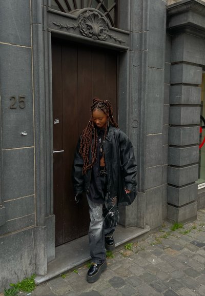 Chaqueta de cuero negra oversized sobre una blusa negra, acompañada de jeans amplios y desgastados. Zapatos negros, múltiples trenzas en el cabello, de pie junto a una puerta de madera.