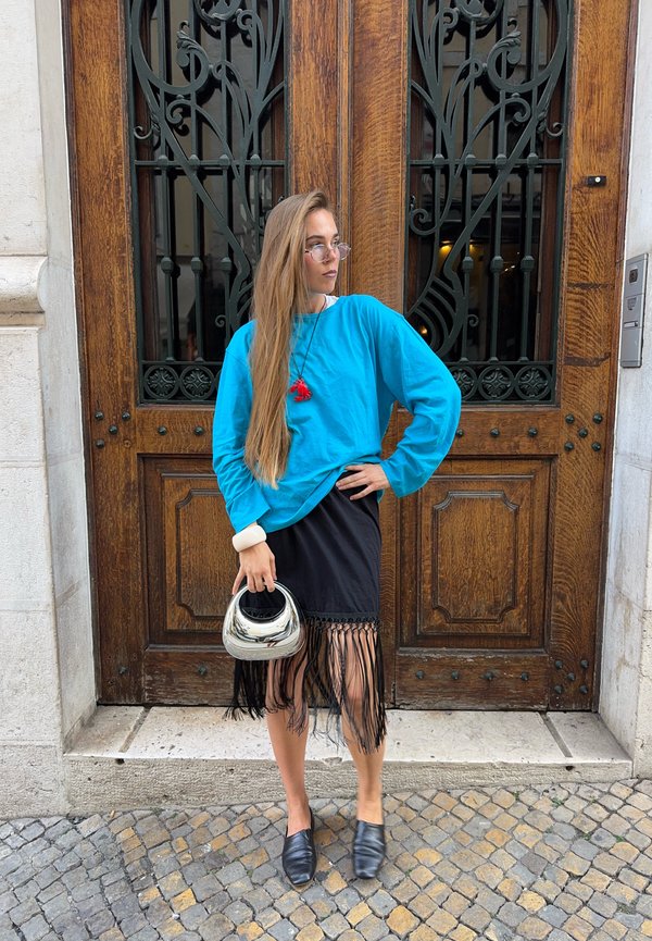 Femme aux longs cheveux portant un pull bleu et une jupe noire à franges, tenant un sac à main argenté, debout devant une porte en bois ornée.