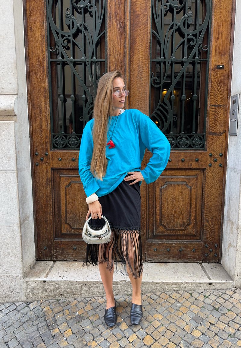 Woman with long hair wearing blue sweater and black fringe skirt, holding silver handbag, standing in front of ornate wooden door.