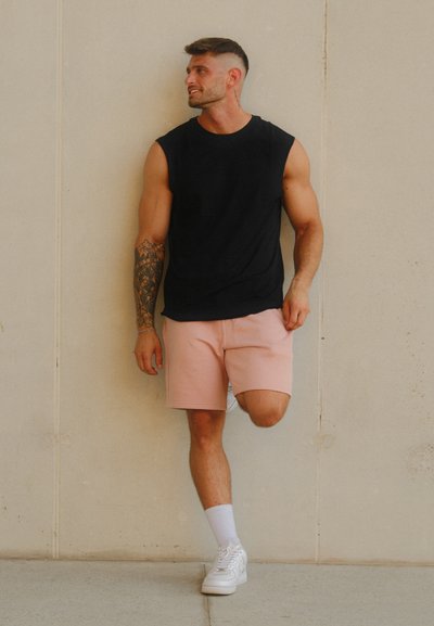 Top negro sin mangas, pantalones cortos rosas, zapatillas blancas y calcetines hasta el tobillo. Piel bronceada y tatuaje visible en el brazo. Pose esbelta contra una pared.