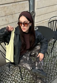 Veste en cuir noire portée sur un haut marron, associée à un pantalon gris à motifs. Un sac fourre-tout jaune clair et des lunettes de soleil brunes surdimensionnées complètent le look.